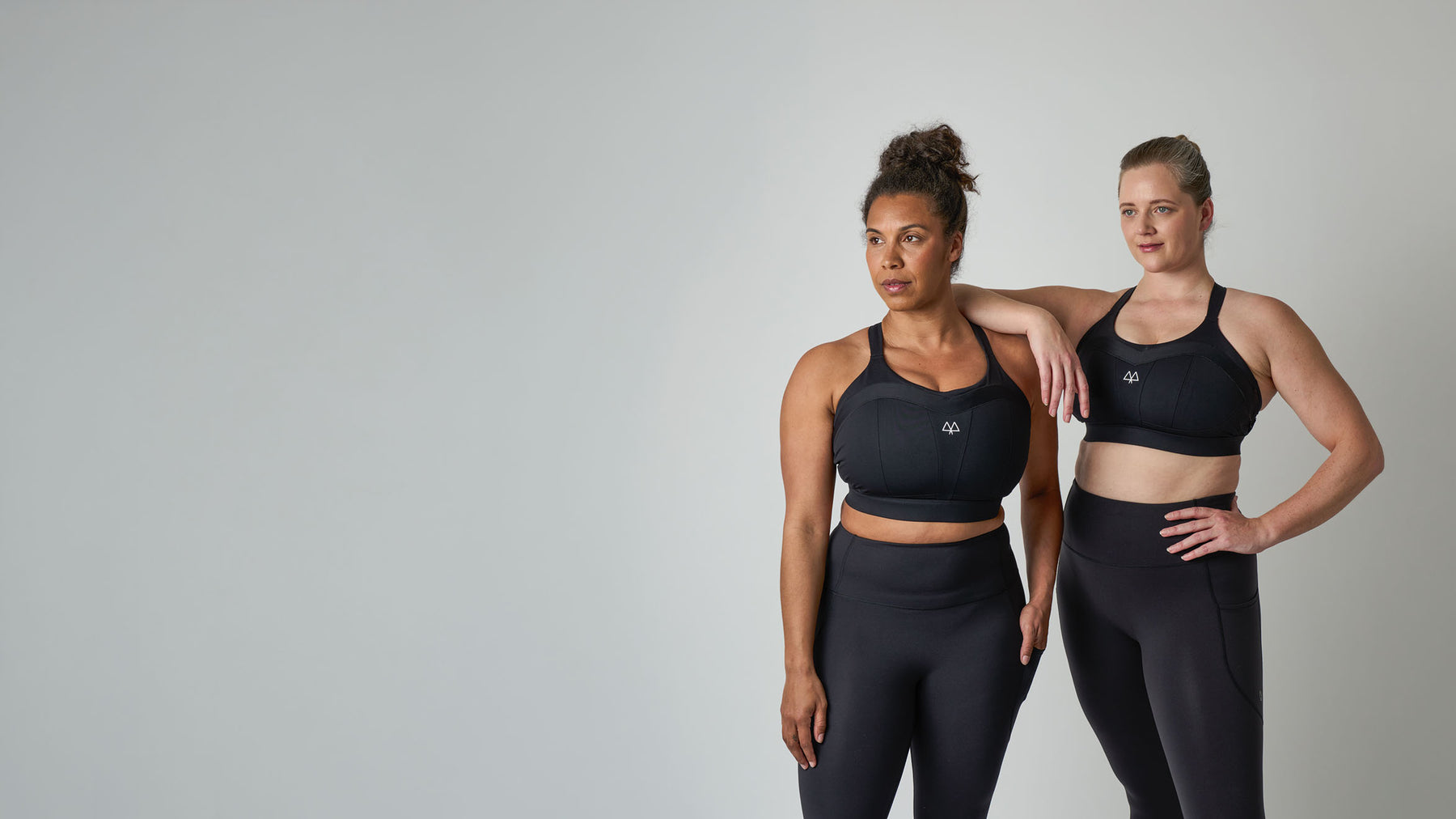Two women modeling matching black athletic sports bra and leggings sets against a plain light gray studio background.