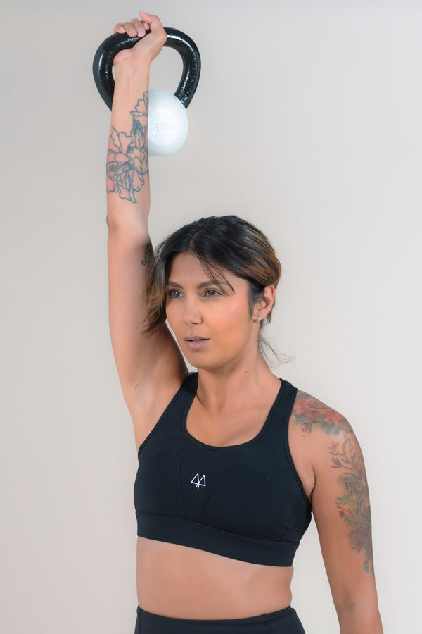 Athlete with tattoos lifting a White Kettlebell above her head while wearing the Empower Sports Bra in Black.